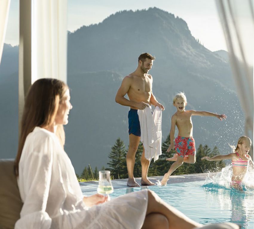 A family at the outdoor pool, the woman on a lounger, the children jumping into the water, the father standing at the edge.
